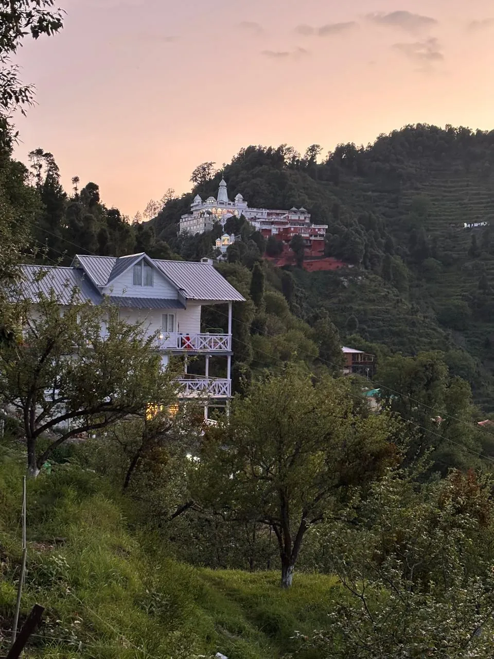 The homestay at dusk with warm lights on and a temple on the ridge above